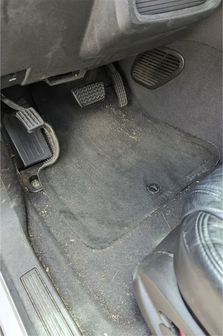 The driver footwell of an SUV before detailing: carpet and floor mat covered in dried grass, dirt and debris, edges caked with grime