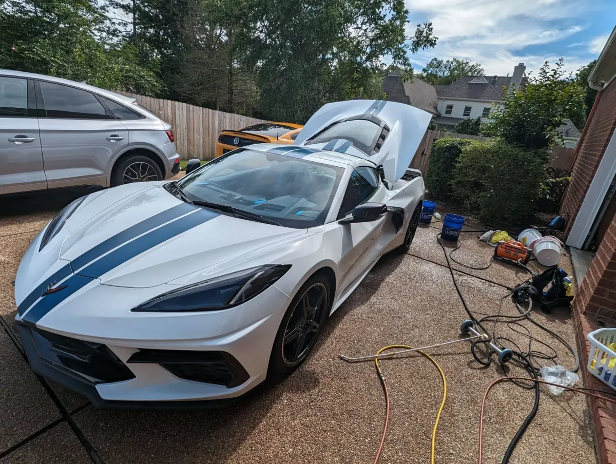 A white mid-engine Corvette with blue stripes on a driveway, hatch open, buckets and pressure washer set up beside it