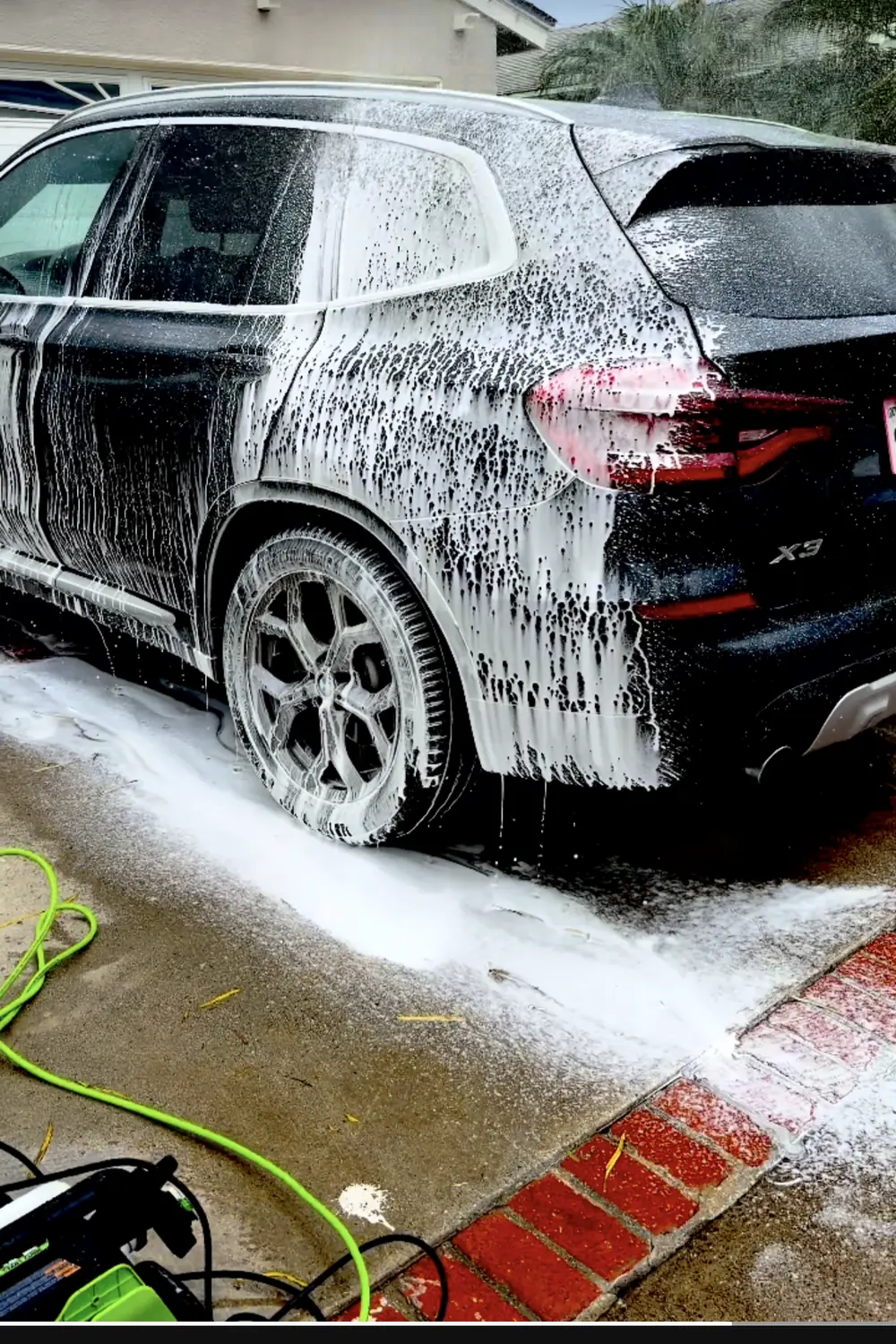 A black BMW X3 under thick white foam in a residential driveway, pressure washer and hose on the concrete