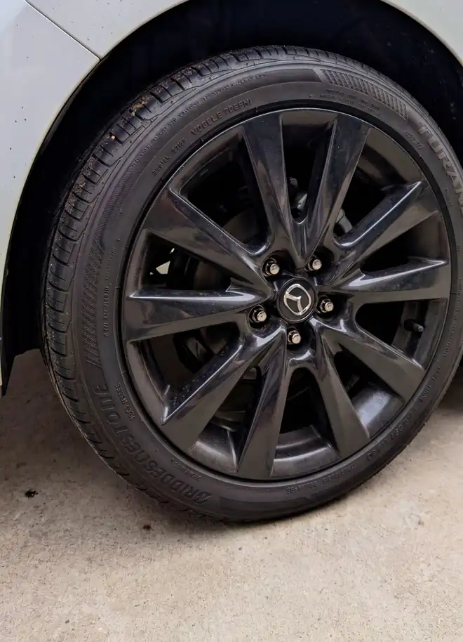 A gloss black alloy wheel and freshly dressed tire on a white Mazda3, photographed in a driveway after detailing